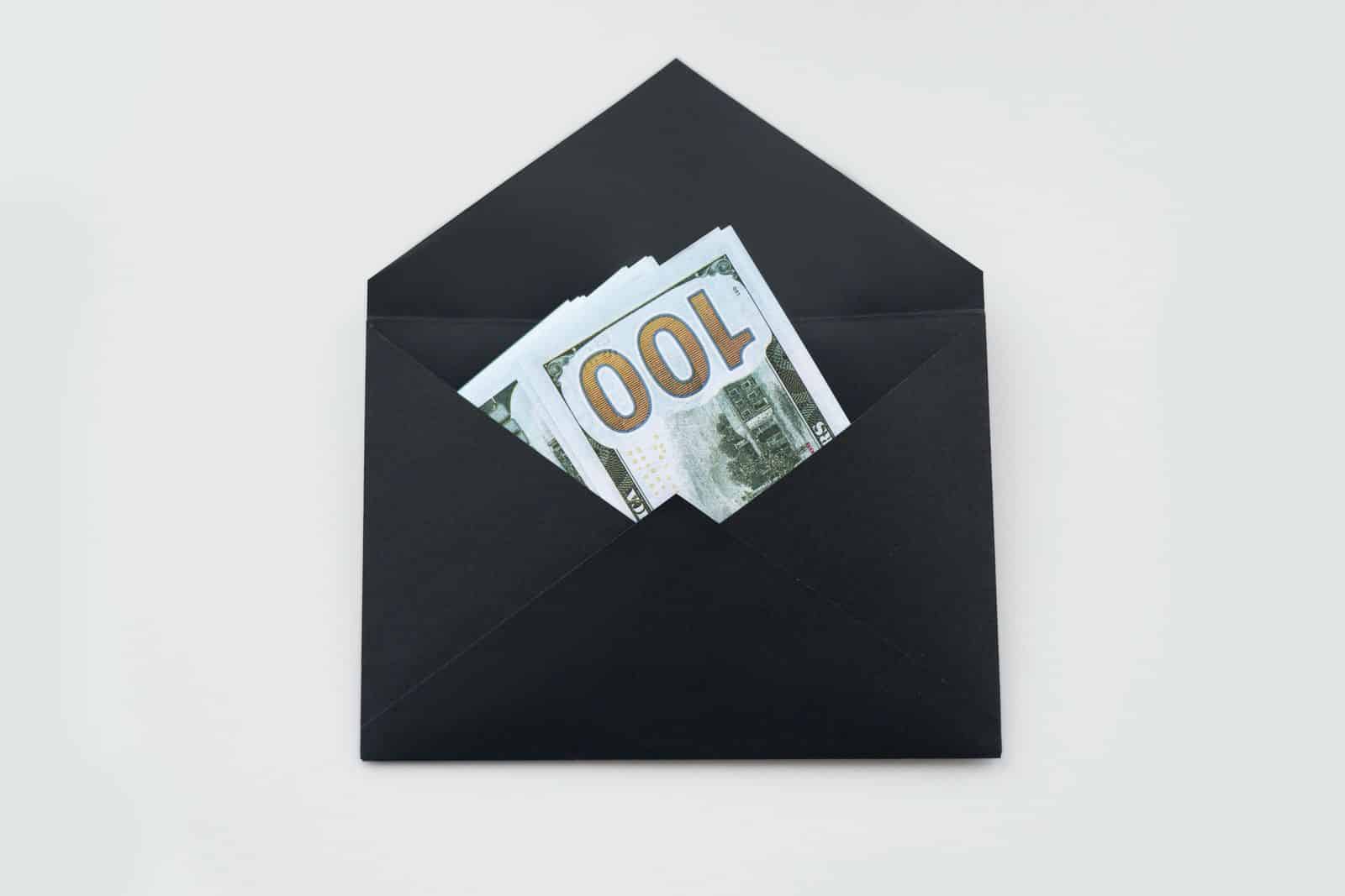 Lehrlingsportal.at Ein schwarzer Umschlag mit mehreren 100-Dollar-Scheinen, fotografiert auf weißem Hintergrund.