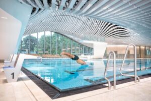 Lehrlingsportal.at Eine Person taucht in ein Hallenbad mit moderner Architektur ein, das sich durch eine wellenförmige Decke und große Fenster mit Blick ins Grüne auszeichnet.