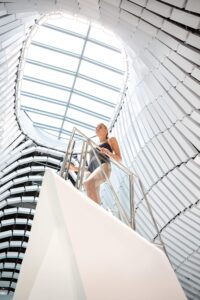 Lehrlingsportal.at Eine Frau in Sportkleidung steht auf einer modernen weißen Treppe unter einem großen, ovalen Oberlicht in einem hellen, futuristischen Gebäude.