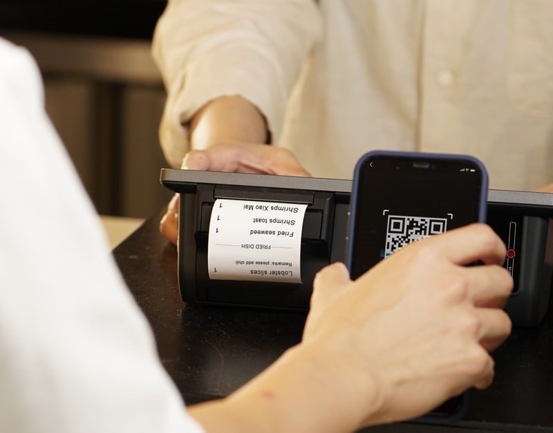 Lehrlingsportal.at Eine Person scannt mit ihrem Smartphone einen QR-Code an einem Zahlungsterminal, während eine andere Person das Gerät ruhig hält.