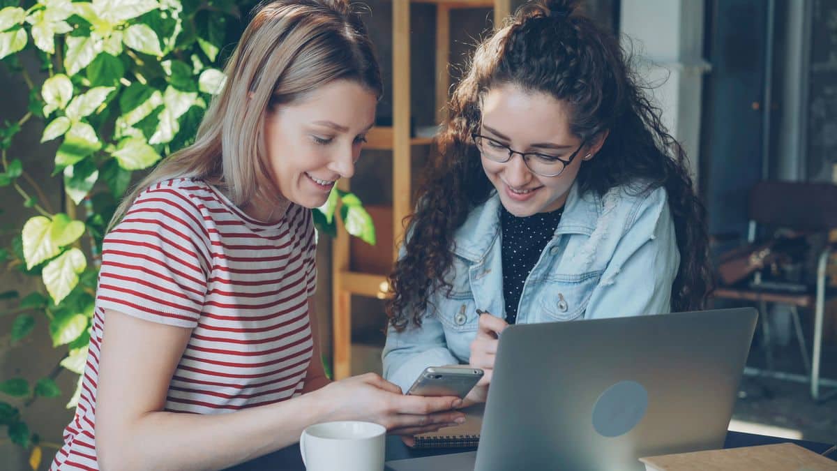 Lehrlingsportal.at Zwei Frauen sitzen an einem Tisch mit einem Laptop, wobei die eine der anderen etwas auf ihrem Handy zeigt. Sie lächeln und scheinen zusammenzuarbeiten oder etwas zu besprechen.