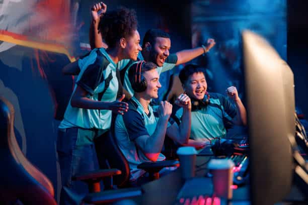 Lehrlingsportal.at Vier junge Erwachsene in einheitlichen Trikots feiern gemeinsam an einer Spielkonsole, lächeln und recken die Arme in die Höhe, während sie in einem schwach beleuchteten Raum auf die Computerbildschirme blicken.