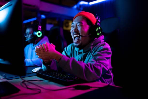 Lehrlingsportal.at Eine Person mit Kopfhörern und roter Mütze sitzt vor einem Computer, lächelt und klatscht in die Hände, umgeben von bunten Lichtern in einer Gaming-Umgebung.