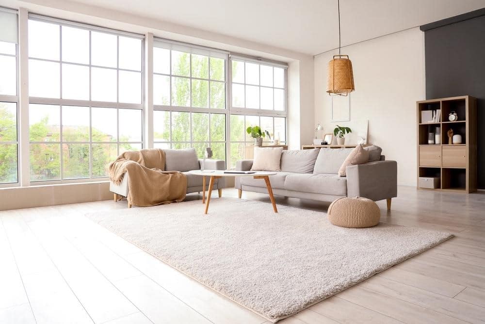 Lehrlingsportal.at Helles Wohnzimmer mit großen Fenstern, hellen Holzböden, einem beigen Sofa und Sessel, einem Couchtisch, einem Teppich sowie einer minimalistischen Einrichtung mit Pflanzen und Regalen.