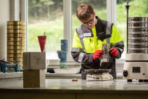 Lehrlingsportal.at Eine Person in Schutzkleidung untersucht Kies mit einem Sieb in einem Labor, auf dessen Tisch wissenschaftliche Geräte stehen.
