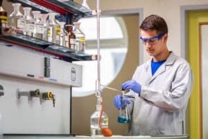 Lehrlingsportal.at Eine Person in einem Laborkittel und mit Schutzbrille gießt in einer Laborumgebung, in der Chemikalien in Regalen stehen, Flüssigkeit aus einem Becherglas in einen Kolben.