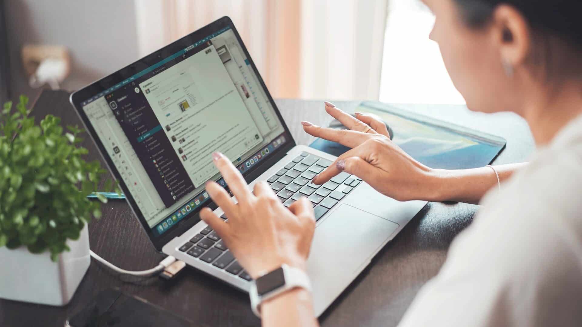 Lehrlingsportal.at Eine Person sitzt an einem Schreibtisch vor einem Laptop und tippt auf der Tastatur, während auf dem Bildschirm Slack geöffnet ist; daneben liegen ein Smartphone, ein Notizbuch und eine kleine Topfpflanze.