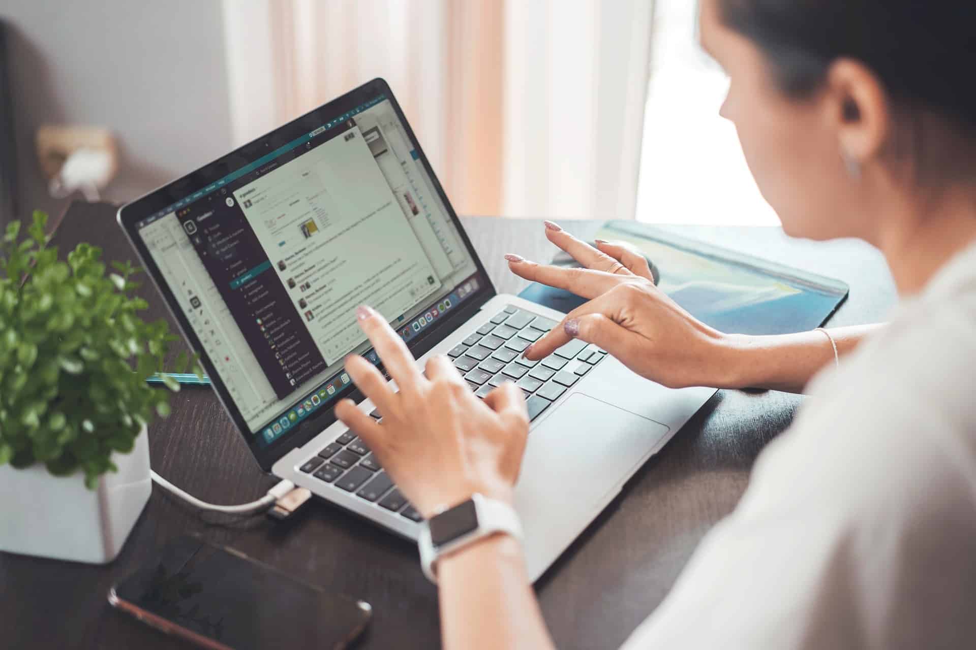 Lehrlingsportal.at Eine Person sitzt an einem Schreibtisch vor einem Laptop und tippt auf der Tastatur, während auf dem Bildschirm Slack geöffnet ist; daneben liegen ein Smartphone, ein Notizbuch und eine kleine Topfpflanze.