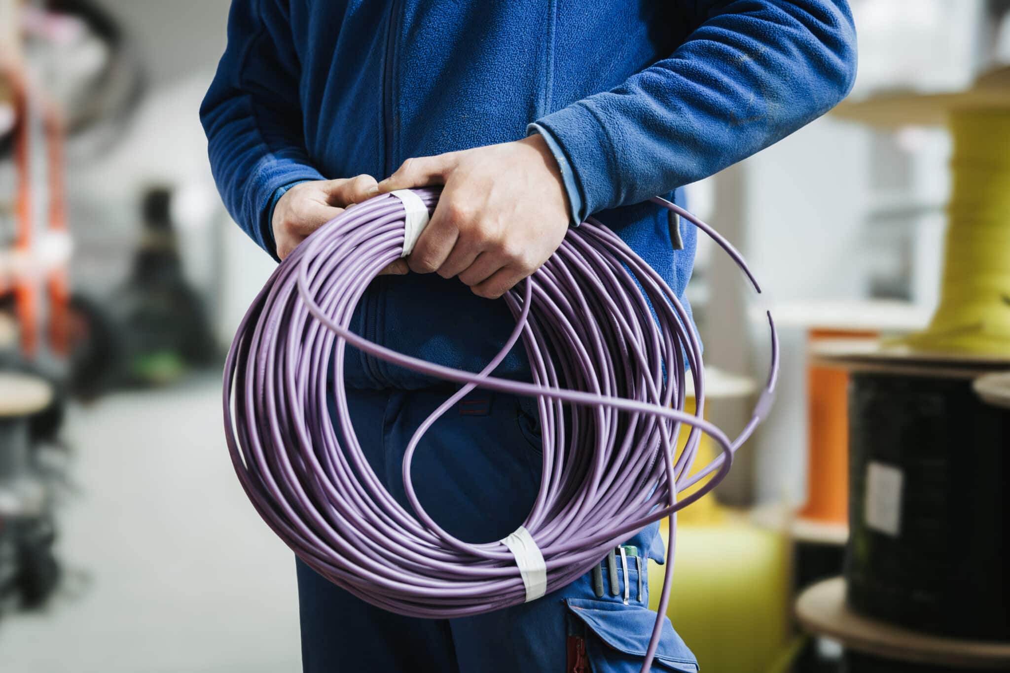 Lehrlingsportal.at Eine Person in einer blauen Jacke, die eine große Rolle lila Kabel in der Hand hält und in einem Innenraum steht, im Hintergrund sind Drahtspulen zu sehen.