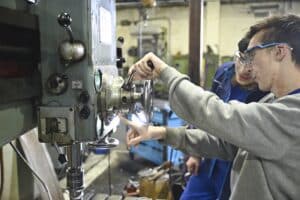 Lehrlingsportal.at Zwei Personen, die Schutzbrillen tragen, bedienen in einer Werkstatt eine große Industriemaschine und konzentrieren sich dabei auf die Einstellung der Steuerelemente.