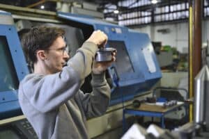 Lehrlingsportal.at Ein Mann in einem grauen Sweatshirt justiert ein Metallteil in einer Fabrikhalle, während er vor blauen Industriemaschinen steht.