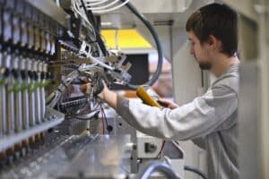 Lehrlingsportal.at Ein Techniker verwendet ein tragbares Diagnosegerät, um die Verkabelung und die Komponenten im Inneren von Industriemaschinen zu prüfen und zu untersuchen.