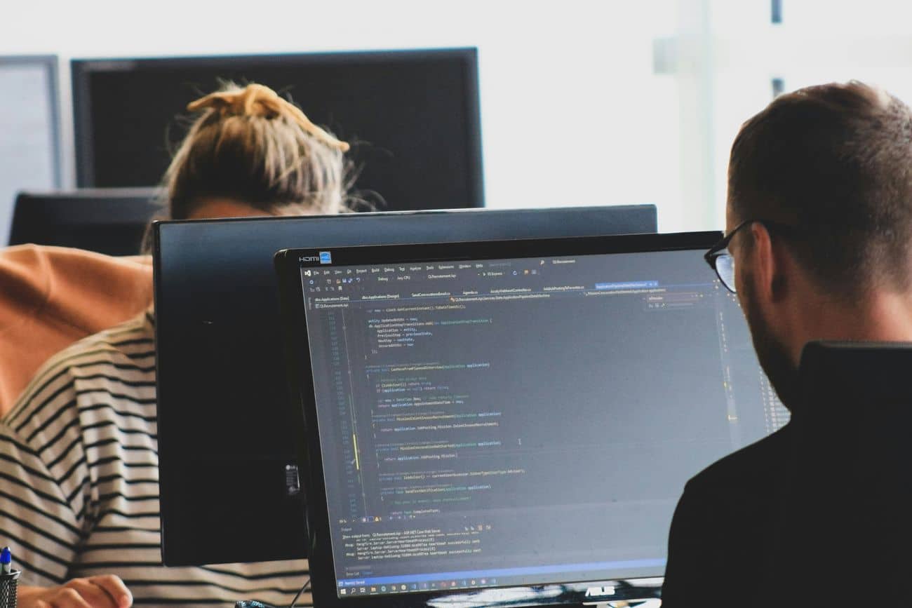 Lehrlingsportal.at Zwei Personen arbeiten an Schreibtischen mit Computerbildschirmen. Auf einem Bildschirm sind Codezeilen zu sehen, was auf Softwareentwicklung oder Programmierarbeiten in einer Büroumgebung hindeutet.