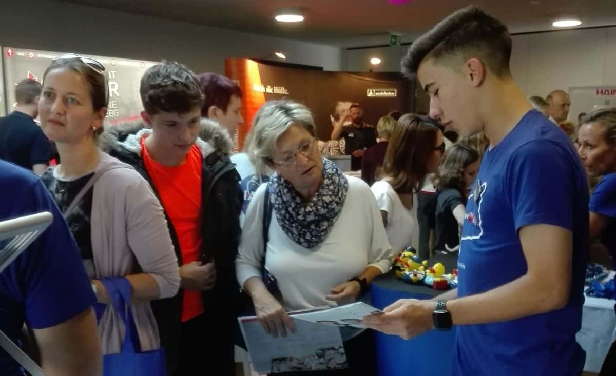 Lehrlingsportal.at Ein junger Mann in einem blauen Hemd unterhält sich mit einer Frau, die Papiere in der Hand hält, bei einer gut besuchten Veranstaltung in einem Saal, in dessen Nähe mehrere Personen stehen.