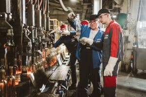 Lehrlingsportal.at Arbeiter in Schutzkleidung prüfen Glasflaschen an einem Fließband in einer Fabrikhalle.