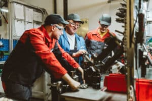 Lehrlingsportal.at Drei Arbeiter in Uniform bedienen Maschinen in einer Industriehalle und konzentrieren sich gemeinsam auf eine Aufgabe in der Metallbearbeitung.