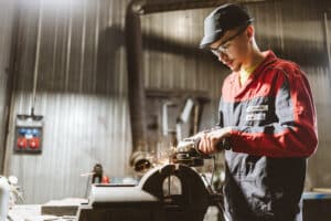 Lehrlingsportal.at Eine Person in einer rot-blauen Uniform bearbeitet in einer Industriehalle mit einem Winkelschleifer einen Metallgegenstand, wobei Funken entstehen.