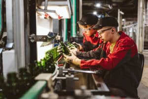 Lehrlingsportal.at Zwei Fabrikarbeiter in roten Uniformen prüfen grüne Glasflaschen an einem Fließband unter grellem Licht in einer Industrieumgebung.