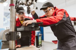 Lehrlingsportal.at Zwei Arbeiter in Schutzkleidung und roten Uniformen bedienen eine Bohrmaschine in einer Industriehalle.