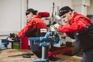 Lehrlingsportal.at Zwei Personen in roten und marineblauen Uniformen arbeiten an Werkbänken in einer Industriewerkstatt mit Metallschraubstöcken und Werkzeugen.
