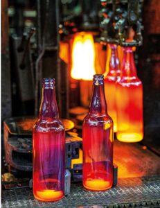 Lehrlingsportal.at In einer Produktionsstätte werden drei glühend heiße Glasflaschen geformt, im Hintergrund ist geschmolzenes Glas zu sehen.