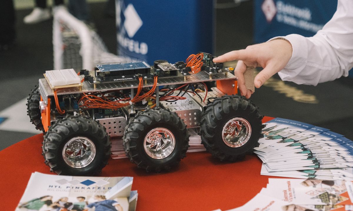 Lehrlingsportal.at Eine Person zeigt bei einer Veranstaltung auf ein elektronisches sechsrädriges Robotermodell, das zusammen mit Broschüren auf einem Tisch ausgestellt ist.