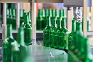 Lehrlingsportal.at Grüne Glasflaschen bewegen sich in einer Abfüllanlage auf einem Förderband, umgeben von Industriemaschinen.