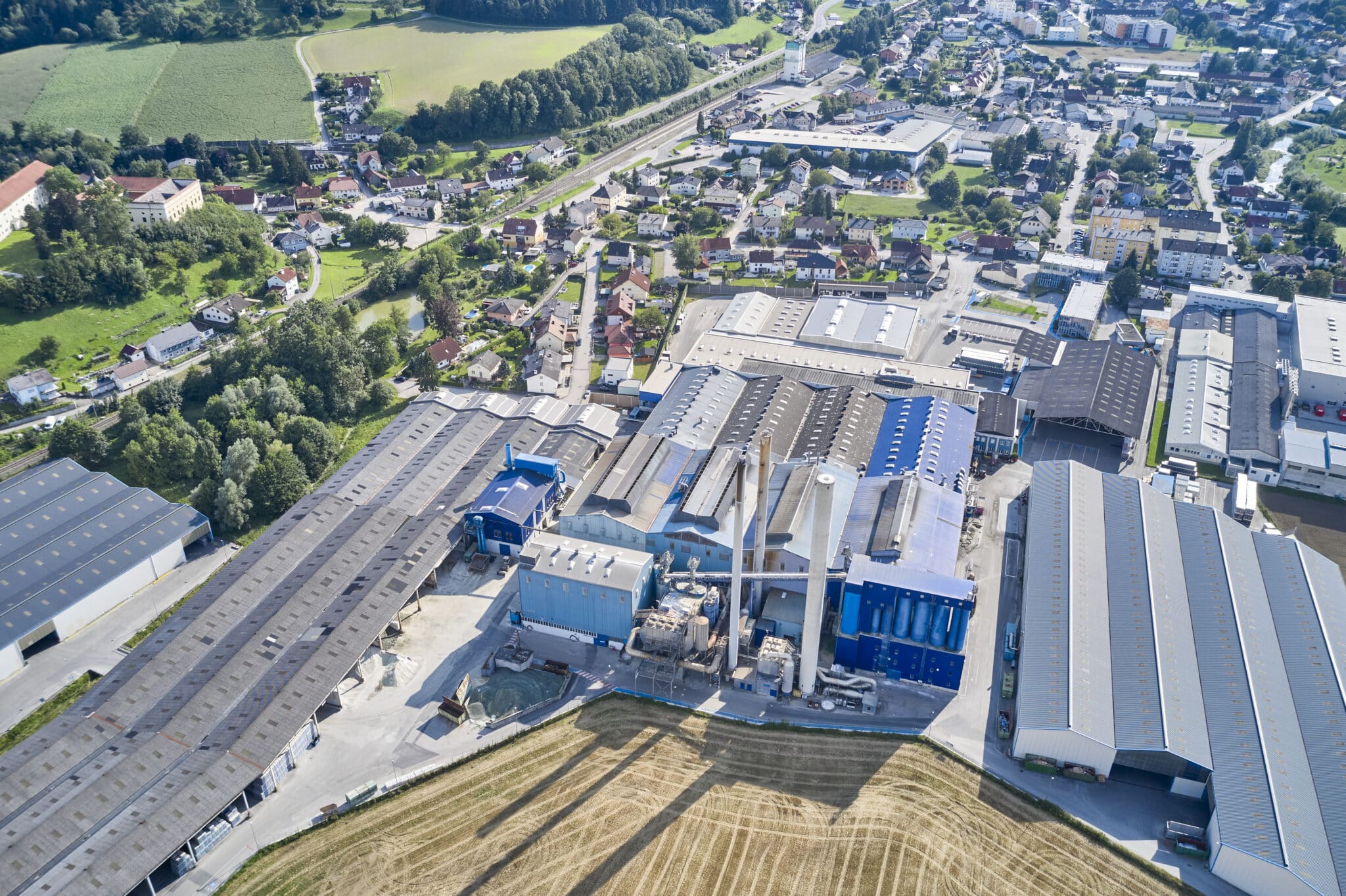 Lehrlingsportal.at Luftaufnahme eines Industriekomplexes mit blauen und grauen Gebäuden, umgeben von Feldern und Wohngebieten unter klarem Himmel.