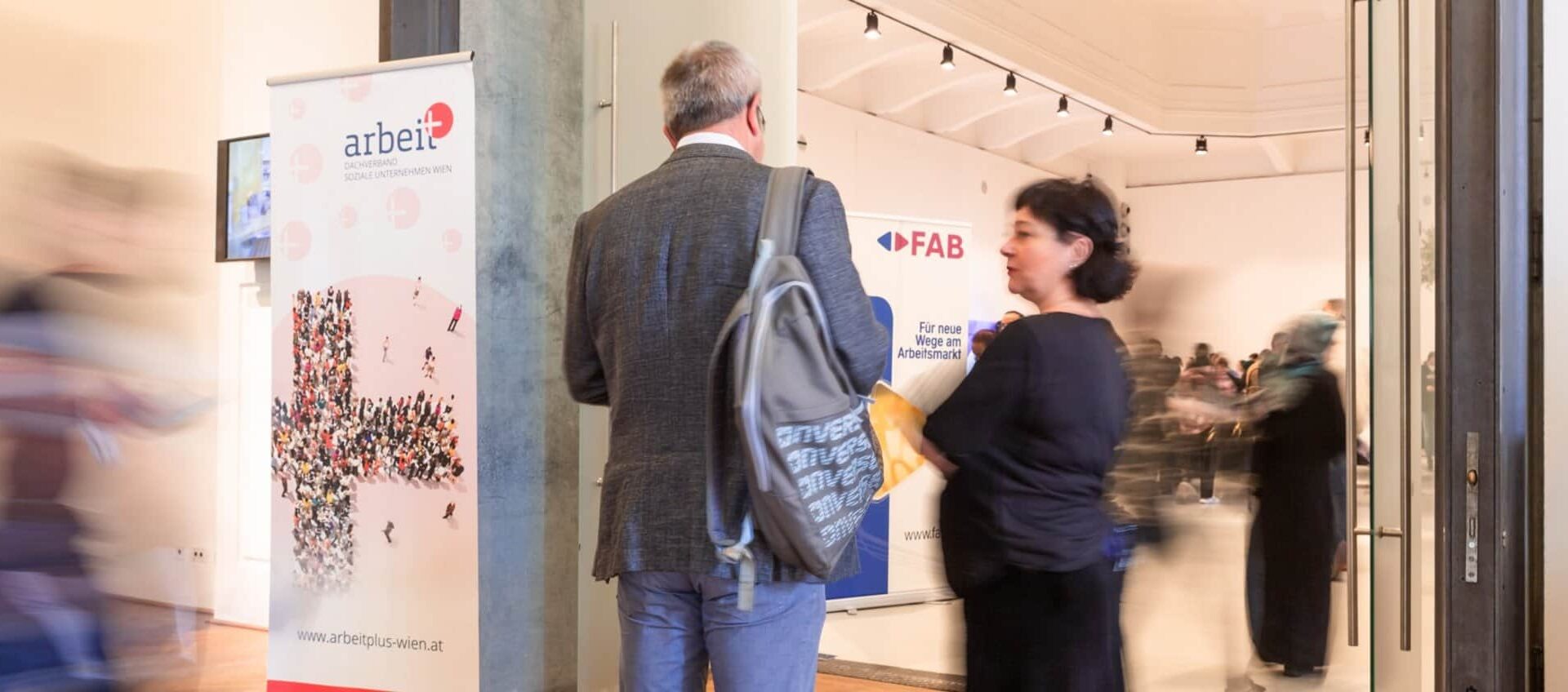 Lehrlingsportal.at Zwei Personen stehen in einem belebten Flur neben einem Banner mit der Aufschrift „arbeit plus“, während im Hintergrund im Inneren des Gebäudes weitere Menschen zu sehen sind, die durch die Bewegung unscharf erscheinen.