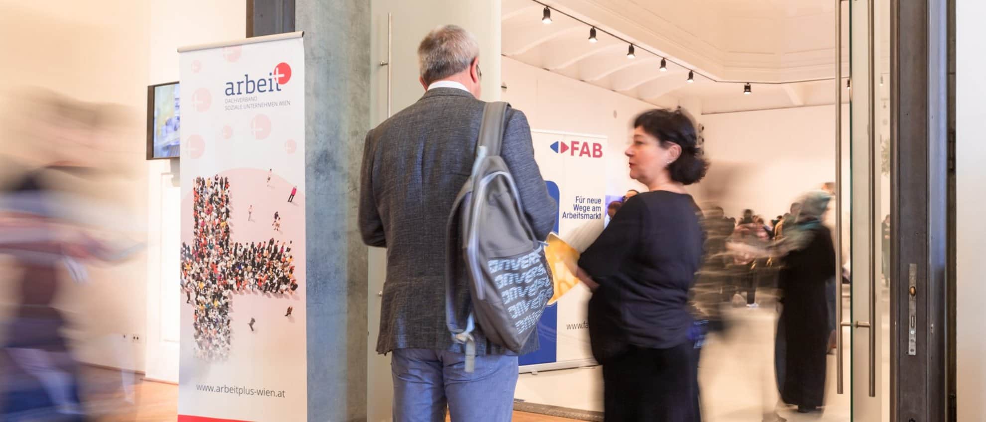 Lehrlingsportal.at Zwei Personen stehen in einem belebten Flur neben einem Banner mit der Aufschrift „arbeit plus“, während im Hintergrund im Inneren des Gebäudes weitere Menschen zu sehen sind, die durch die Bewegung unscharf erscheinen.