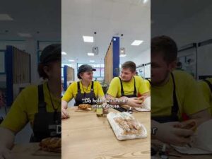 Lehrlingsportal.at Zwei Personen in gelben Hemden und Schürzen sitzen an einem Tisch in einer Cafeteria und essen; vor ihnen stehen Brot und Snacks. Auf dem Bild sind deutsche Untertitel zu sehen.