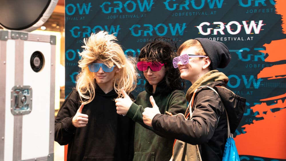 Lehrlingsportal.at Drei Personen mit Perücken und bunten Shutter-Shades posieren mit erhobenem Daumen vor einer blau-orangefarbenen Kulisse des „GROW“-Jobfestivals.
