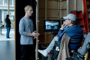 Lehrlingsportal.at Zwei Männer unterhalten sich am Filmset; der eine steht und gestikuliert, der andere sitzt auf einem Stuhl neben einem Monitor, auf dem eine Szene zu sehen ist. Im Hintergrund sind weitere Mitglieder der Filmcrew und Ausrüstung zu sehen.