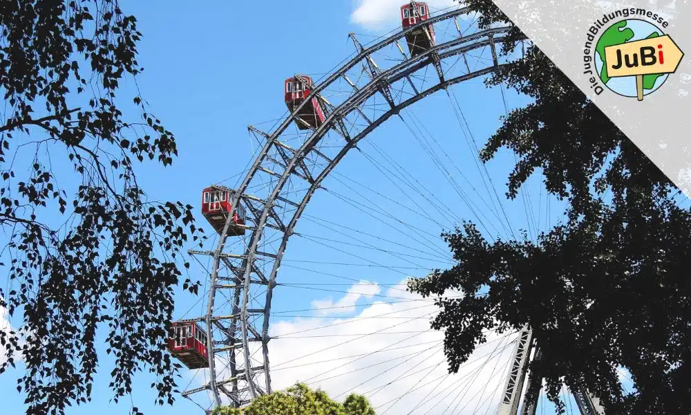 Lehrlingsportal.at Ein großes Riesenrad mit roten Gondeln vor blauem Himmel, teilweise von Ästen eingerahmt; in der rechten oberen Ecke ist ein JuBi-Logo zu sehen.