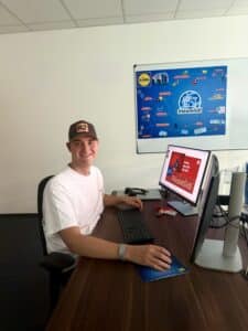 Lehrlingsportal.at Ein junger Mann mit einer Baseballkappe sitzt an einem Schreibtisch vor einem Computer, hinter ihm hängt ein Lidl-Organigramm an der Wand.