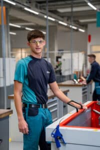 Lehrlingsportal.at Ein junger Mann mit Schutzbrille und blauer Arbeitskleidung steht in einer Werkstatt und stützt seine Hand auf eine Maschine.