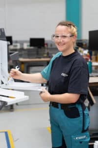 Lehrlingsportal.at Eine lächelnde Person mit Schutzbrille und in Arbeitskleidung justiert in einer Werkstatt mit einem Schraubenschlüssel einen Metallrahmen.