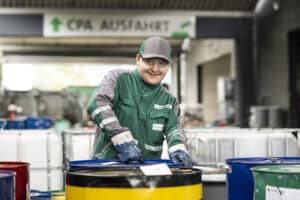 Lehrlingsportal.at Ein Lagerarbeiter in grüner Uniform, mit Mütze und Schutzbrille, der mit Industriefässern arbeitet.