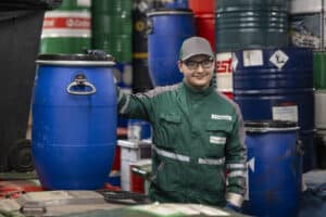Lehrlingsportal.at Ein Arbeiter in grüner Uniform und mit grüner Mütze steht in einem Lagerraum neben einem großen blauen Industriefass.