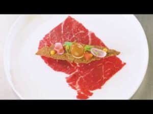 Lehrlingsportal.at Dünne Scheiben rohes Rindfleisch, belegt mit einem knusprigen Cracker, einem Eigelb, Radieschenscheiben, Blattgemüse und bunten Soßen, angerichtet auf einem weißen Teller.