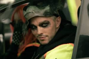 Lehrlingsportal.at Auf dem Fahrersitz sitzt eine Person mit einer Wolfsmaske und dunklem Augen-Make-up, auf dem Beifahrersitz eine Person mit pinkfarbenen Haaren.