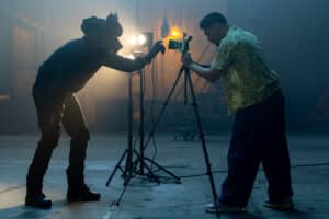 Lehrlingsportal.at Eine Person mit einer Werwolfmaske stellt eine Studiolampe ein, während ein Filmemacher eine Kamera auf einem Stativ bedient.