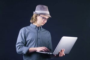 Bursche mit Hut arbeitet im Stehen am Laptop vor schwarzem Hintergrund