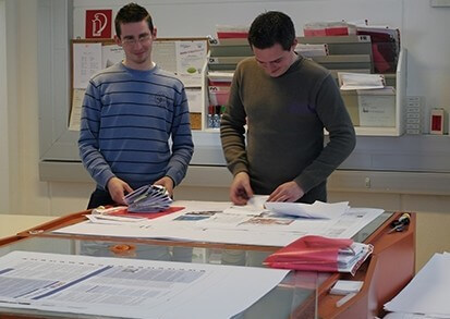 Druckerei Boesmueller Zwei Mitarbeiter Stehen Vor Tisch Pruefen Druckmittel