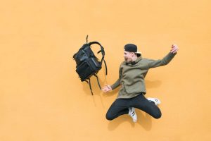 Teenager in olivgrünem Sweatshirt und Käppi macht Luftsprung und wirft Rucksack vor orangenem Hintergrund