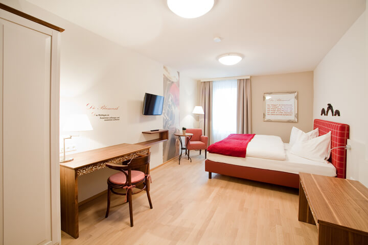 Hotelzimmer mit rotem Doppelbett, Schreibtisch und TV